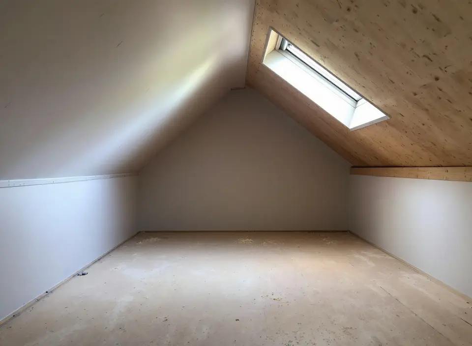 Een lege zolder in afwerking met lage knieschotten van witte gipsplaat, een houten dakbeschot met een dakraam en wat zaagsel op de vloer