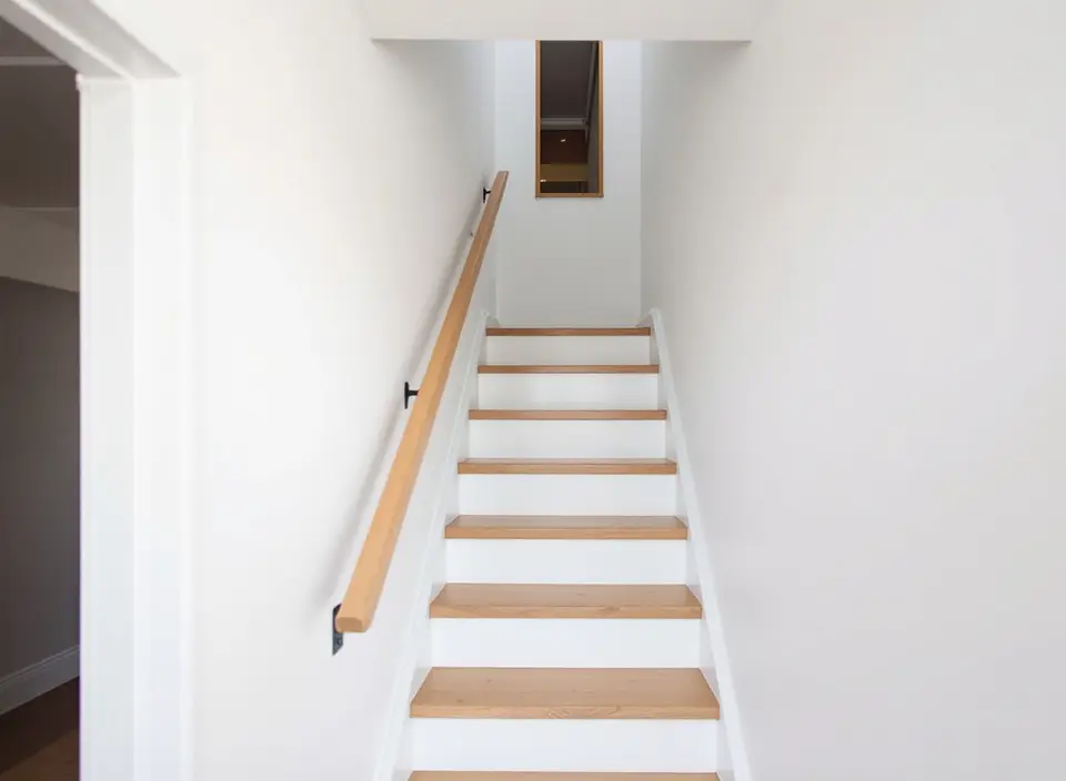 Een rechte, wit geschilderde vaste trap met eikenhouten treden en een houten leuning aan de muur, die naar het trapgat van de zolder leidt